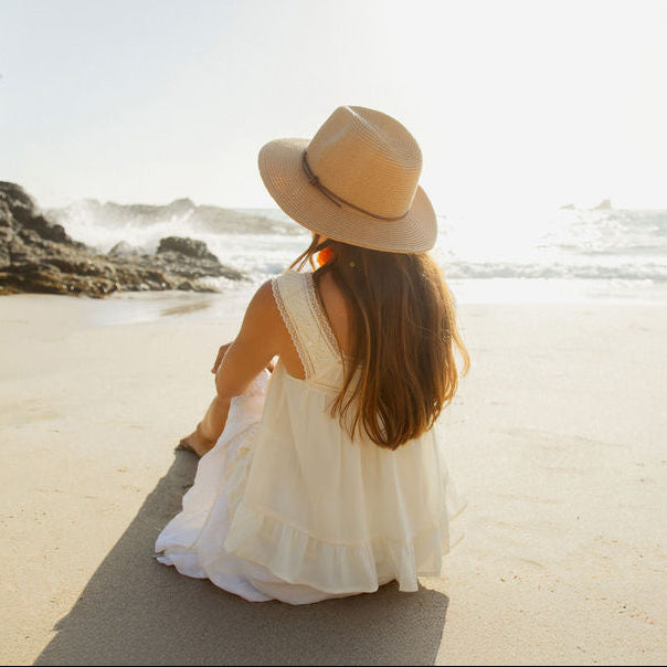 Woman in a white dress and hat sitting on a beach with ocean and rocks in the background